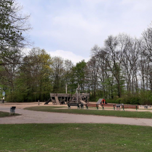 Das Bild zeigt den Spielplatz in der Rheinaue aus einigen Metern Entfernung. In der Mitte ist das große Spielschiff aus Holz zu sehen. Um dieses herum befindet sich eine große Sandfläche. Im Hintergrund stehen Bäume.
