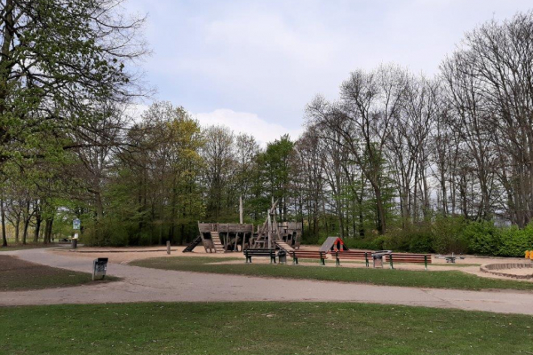 Das Bild zeigt den Spielplatz in der Rheinaue aus einigen Metern Entfernung. In der Mitte ist das große Spielschiff aus Holz zu sehen. Um dieses herum befindet sich eine große Sandfläche. Im Hintergrund stehen Bäume.
