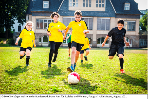 Sechs Kinder und Jugendliche mit und ohne Behinderung spielen auf einer Wiese Fußball