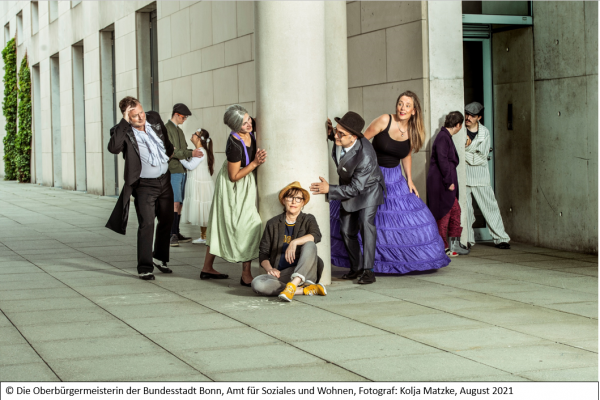 Neun unterschiedliche Menschen auf einem überdachten Platz am Kunstmuseum. Die Menschen sind Schauspielende in einer inklusiven Theatergruppe.