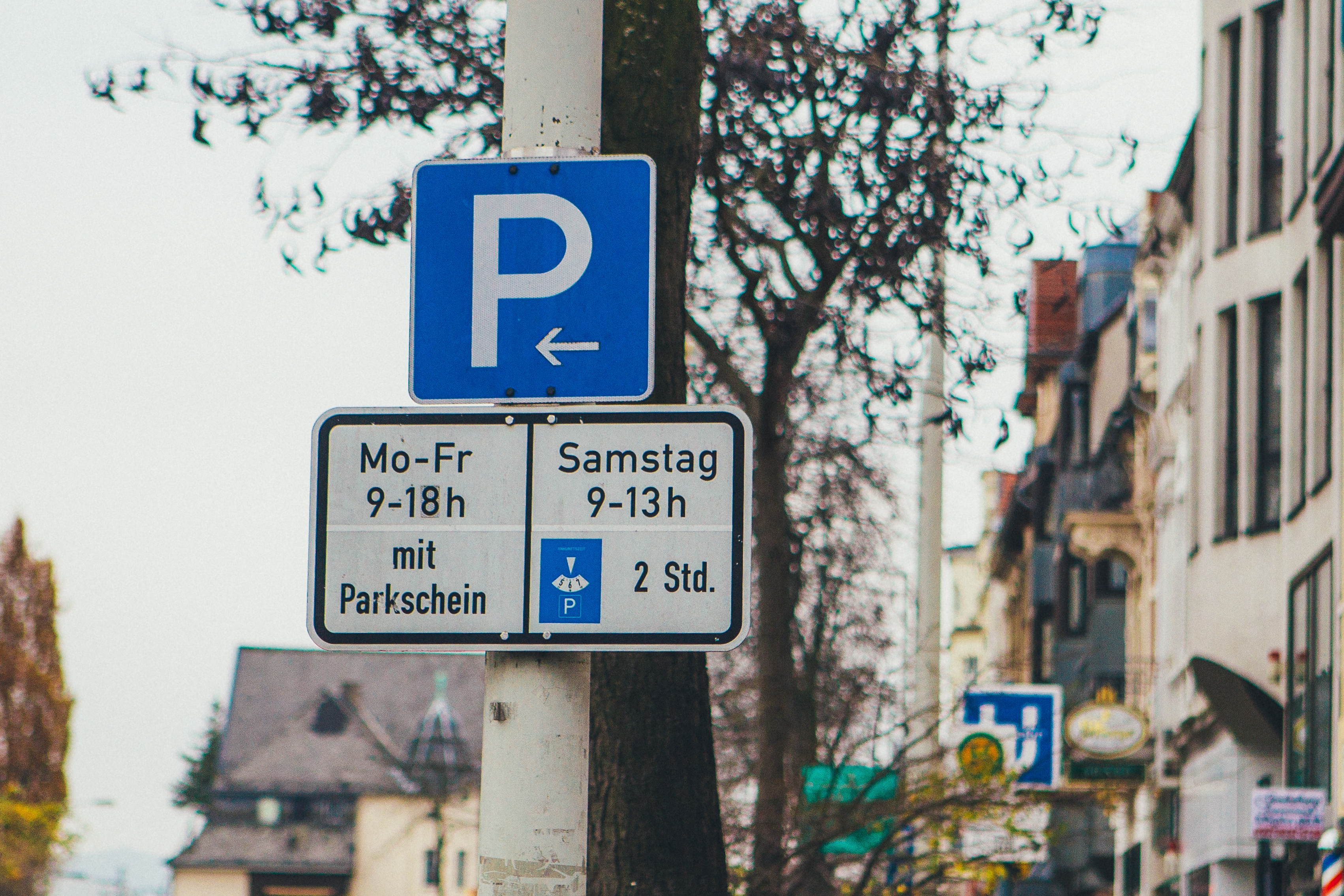 An einer Laterne hängt ein blaues Parken Schild, mit Mo-Fr von 9-18 Uhr mit Parkschein und Samstags von 9-13 Uhr für 2h mit Parkscheibe. 